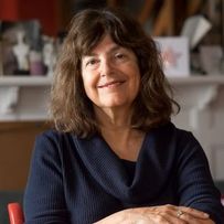 Frau mit schulterlangem gewelltem Haar trägt einen dunkelblauen Pullover mit weitem Kragen und sitzt mit verschränkten Armen vor einem Kamin mit Dekorationen.