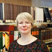 Person mit kurzem blondem Haar, Ohrringen und mehrreihiger Kette vor einem Bücherregal mit vielen Büchern